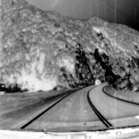 Grayscale frame-camera image of the same daylight scene, captured simultaneously: hillside, road and lane markings all legible.