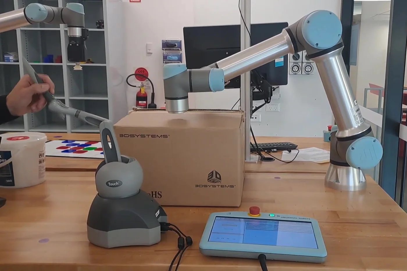 A hand holding the stylus of a 3D Systems Touch haptic device on a workbench, with a Universal Robots UR5e arm posed behind it and the teach pendant in front.